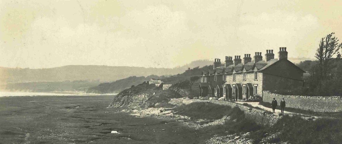 Black and white photo of a terrace of six stone houses facing the sea. Two men are standing in the narrow lane that runs in front of the houses beside the water.