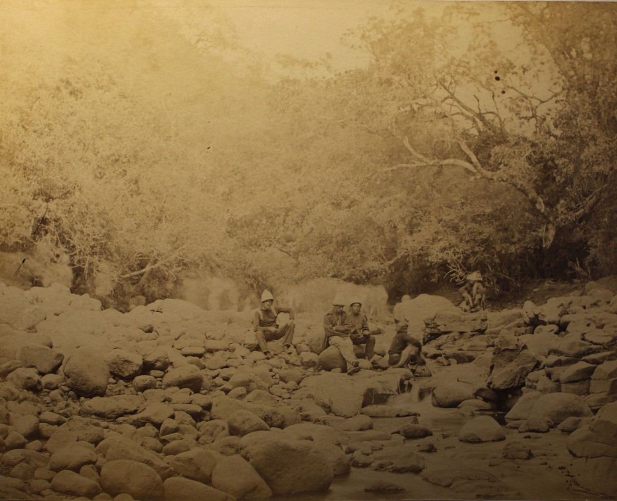 photo of several soldiers sitting on rocks in a dry riverbed surrounded by trees.