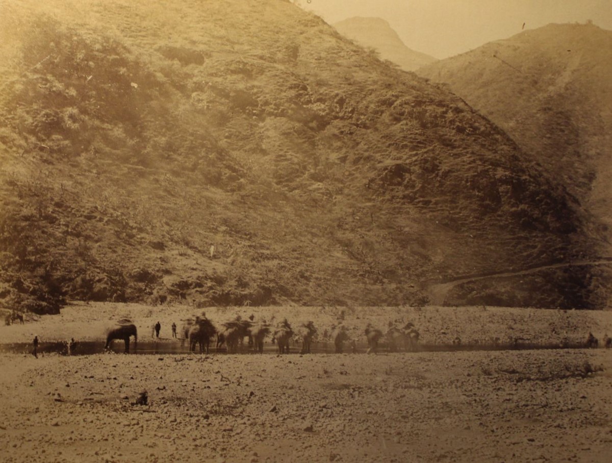 photo of soldiers and mules drinking from a river in a rocky landscape at the foot of steep hills.