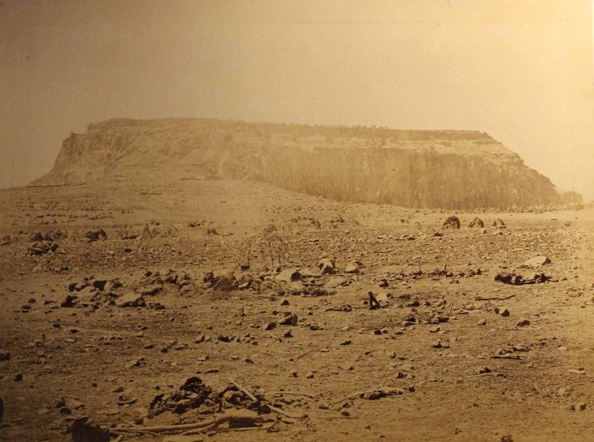 photo of a large hill with a flat top and almost vertical sides. There are buildings on top. The foreground is rocky and barren.