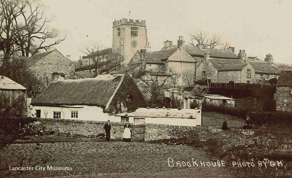 The black and white photo of Brookhouse was taken around 1900. It shows several houses with the church rising above them. The church is perched on a rise. It is made of stone with a square tower with crenellations. At the top of the tower are what look like windows on each side, but they are slatted and are there to allow the sound from the bells to come out. There is a round clock below the west facing one, which is also the one facing the camera. In the foreground of the image is a field and a man and a woman standing by a gate in a waist-height stone wall. The wall leads to a low stone white-washed house. They look to be working people, the man is dressed in black and the woman has a long dark dress or top and skirt and a white apron. The apron starts at her waist and falls almost to the bottom of her skirt.