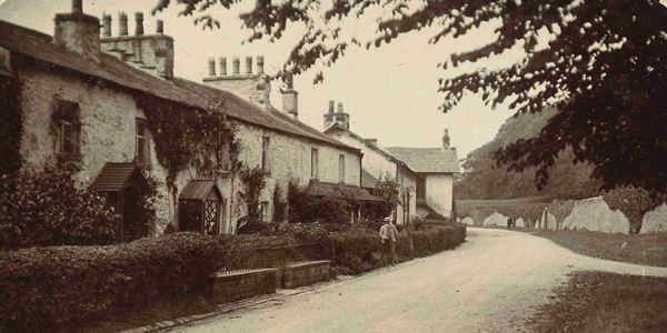 Black and white photo of cottages lining the street in Nether Burrow, looking north towards Over Burrow. There are roses in the front gardens of the cottages and climbing up the walls. A tree overhangs the road and ivy covers a high stone wall on the far side of the street in the background.
