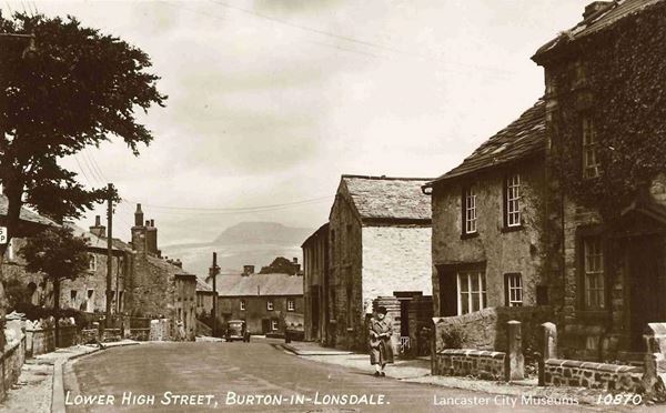 The image shows the High Street of Burton-in-Lonsdale. In the distance is the mountain-fort of Ingleborough with its distinctive flat top. In the foreground on the right is a lady in a coat and black hat. The coat is below the knee and part way down the shin as is typical in the middle of the twentieth century. She is carrying what looks like a letter in her hands. At the end of the street behind her is a black motor car, again looking like a car from the middle of the twentieth century, sturdy with a high roof.