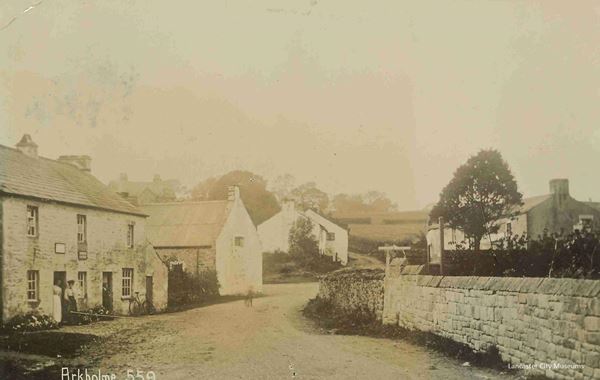 Sepia photo of the road winding between stone cottages and walls in Arkholme. Two women in Edwardian dress watch from a doorway to the left. A bicycle leans against the cottage wall. In the background are fields and trees on a low hill.