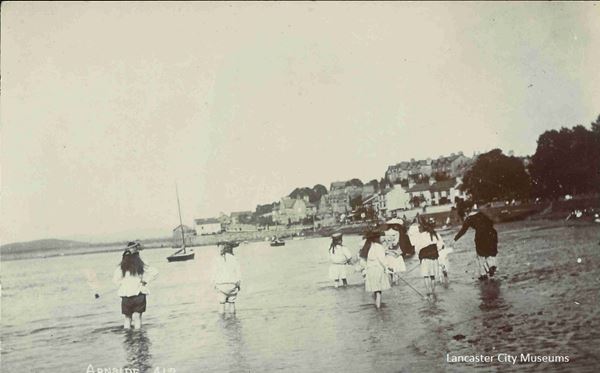 Black and white photo of children in Edwardian dress paddling and playing on the beach, with the houses of Arnside in the background. Several small boats are moored nearby.