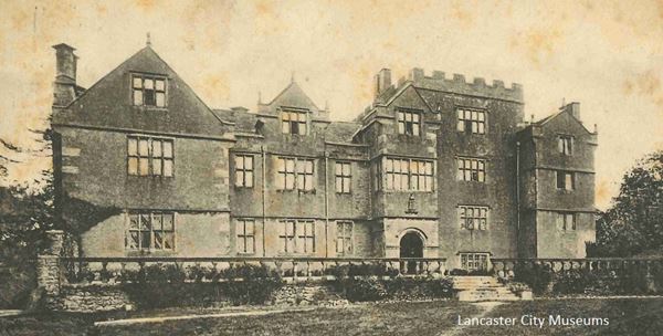 This is an image of Borwick Hall. Borwick Hall is a large building of three stories high and seven large windows across the front at first floor level. The roofline is sometimes pointed and sometimes crenellated, making it look quite higgledy-piggledy. The Hall mainly dates from the 1500s and 1600s and the windows are mullioned. The Hall is built of a dark coloured stone.