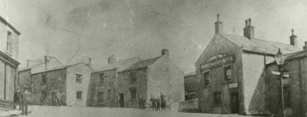 Black and white photo of stone houses in Carnforth. Several men and boys in Victorian clothes are standing around in the street. One building to the right has a large sign reading 'Wines and Spirits'.