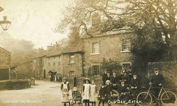 The photo shows a group of Victorian or Edwardian men, one woman and children standing by an old oak tree to the right. Behind them runs a lane with houses. The woman and girls wear white pinafores and the man to the far right has a bicycle. The woman has a pram. The men and boys wear caps.