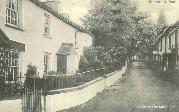 The black and white photo shows a road rising up a hill with stone cottages on both sides. The cottage on the left has been plastered and painted white. The wall in front of it has also been plastered and painted white. The scene looks very pretty.