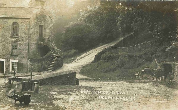 The black and white photo shows the road leading steeply down to a three storey stone mill building dating from the 1800s. The mill has stone steps at the side leading up to a first floor doorway.