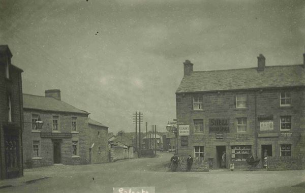 Black and white photo of the main road in Galgate. Two men lean against a low stone wall in front of a stone building with a large 'Shell' sign on the front.