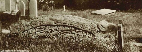The black and white photo shows the Heysham hogback dating from the 900s. It is a piece of stone sculpture about the size of a gravestone and may well have been a grave marker. It is hump-backed with decorative carving depicting scenes and what are thought to be bears grasping each end.
