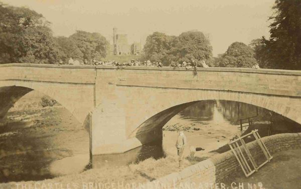 The photograph shows the bridge over the River Wenning at Hornby with Hornby Castle in the background. There is a large group of children on the bridge. The photo dates from around 1900.