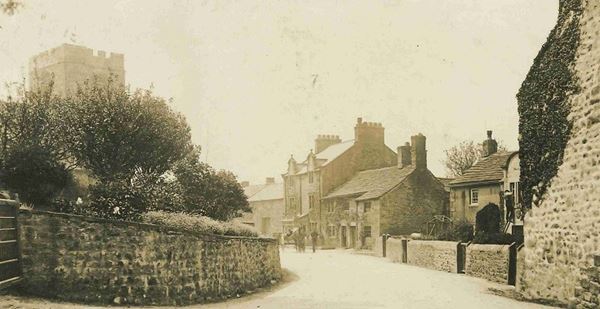 Black and white photo of stone houses and cottages lining the street in Melling. The square church tower rises behind some bushes growing on an earthen mound that was once the motte..