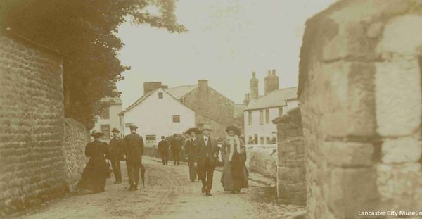 This photo shows men and women in edwardian clothing and hats strolling along the street between high stone walls. In the background are several stone houses, some of them painted white. One has unusually tall chimneys.