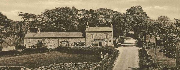 Black and white photo of a long stone building with a sign reading 'Temperance Hotel'. It stands in front of trees and beside a country lane lined with dry stone walls. 