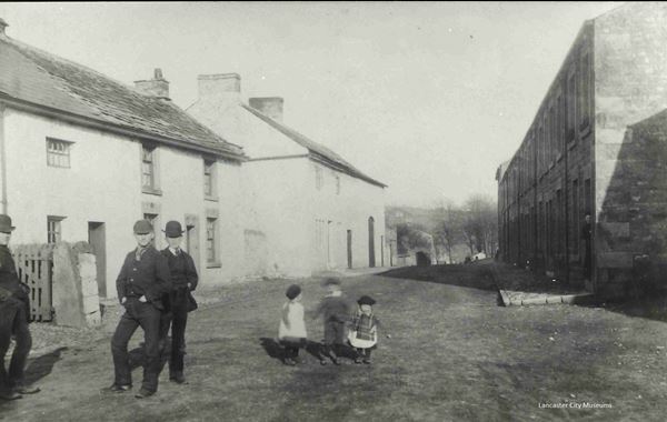 Black and white photo looking along a street with a row of terraced stone houses on the right, and older white-painted cottages on the left. Three young men standing on the left are posing for the photo, while three small children play in the middle of the otherwise empty street.