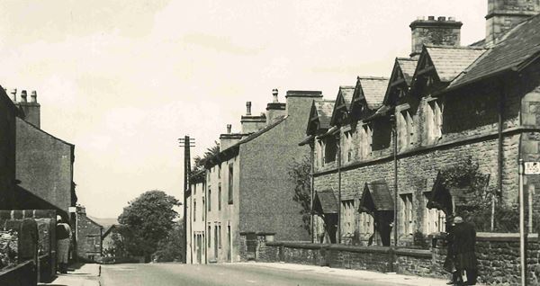 Black and white photo of a street sloping gently downwards away from the camera. Stone houses in various styles line both sides of the road.
