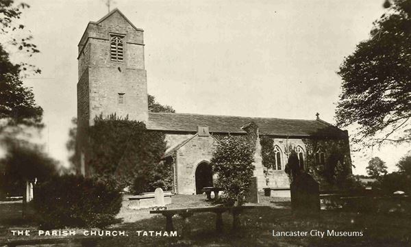 Black and white postcard showing the parish church at Tatham. It's a small stone church with a square tower. The churchyard around it has a number of gravestones and simple stone tombs standing amongst scattered bushes and trees.