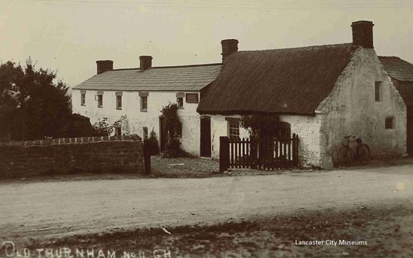 Black and white photo of white painted cottages. The nearest one is a low thatched building. A row of newer slate-roofed cottages has been added on to the side of it.