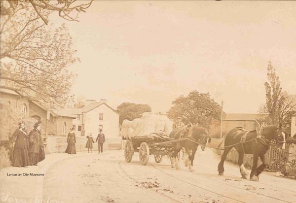 Sepia photo looking along a cobbled street, with trees and stone houses set back on either side. Half a dozen people are walking along the pavement. They're in Victorian dress, mostly women in long skirts, coats and hats or bonnets. A man with a hat is holding a small child by the hand. A wagon heavily laden with sacks is passing by in the road, drawn by two large shire horses.