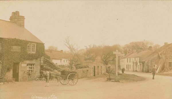 This photo shows the centre of the village of Wennington. The road curves past stone walls and cottages. A signpost with arms pointing in both directions stands beside is a water pump and stone trough. The building to the left is covered in ivy and there is a bicycle leaning against it. In front of the house two men are sitting in a horse-drawn chaise.