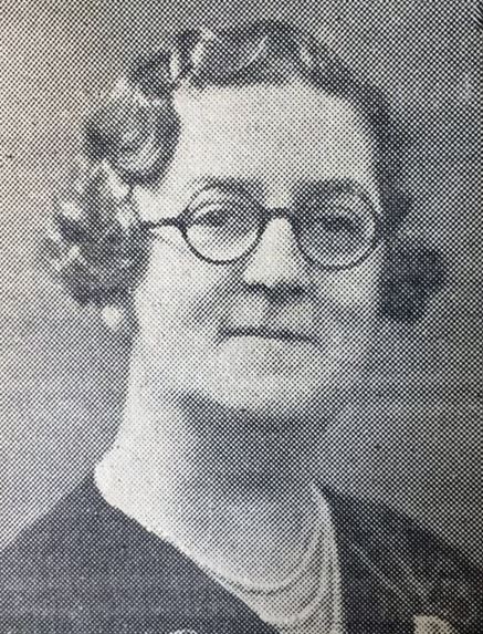 Photo of Muriel Dowbiggin - she has dark hair in a short wavy style and round glasses, earrings, and a necklace with several strings of pearls.