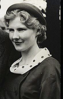 Photo of Noreen Murray. She has blonde hair in a short curled style and wears makeup and stylish clothes: a dark double breasted jacket over a polka dot blouse, and a small hat.