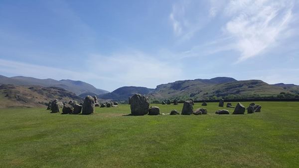 Photo of a complete circle of stones of various sizes and shapes on a flat green field, with a view of the high hills of the lake district in the background.