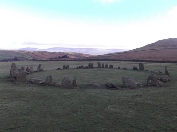 Photo of a complete circle of stones of various sizes and shapes on a flat green field, with a view of the high hills of the lake district in the background.