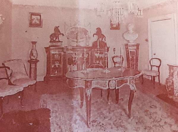 Photo of a room containing elaborate Victorian furniture: a table, chairs, large dresser and several large ornaments.