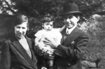 Black and white photo of a woman in a smart jacket standing beside a man in a bowler hat and suit jacket, who is holding a small child. They're both smiling and it seems to be a sunny summer's day. There are trees and bushes in the background.