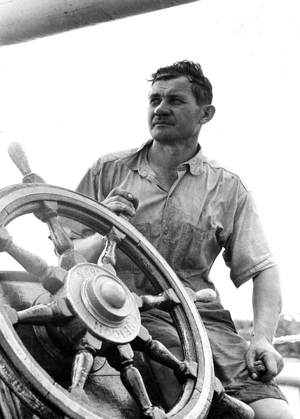 Photo of a rugged and windswept man posing beside a wooden ship's wheel. He's dressed for hot weather in shorts and a rumpled short sleeved shirt.