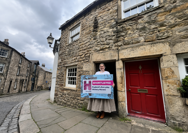 photo of woman in georgian costume standing in front of stone cottage, holding a placard about the everyday history heroes award