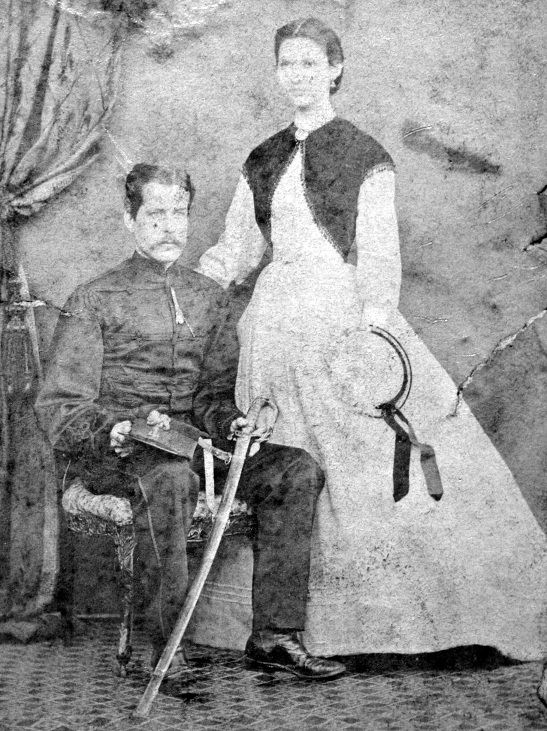 studio photo of a seated man in uniform and a woman standing beside him with a hand on his shoulder, wearing a full-skirted victorian dress.